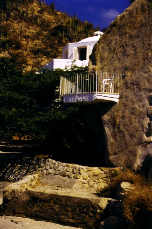 Las blancas masas y formas de los volúmenes contrastan con lo agreste del paisaje. La memoria de la arquitectura del Mediterráneo, evidente en este caso, acentúa el contrapunto entre las formas, colores y texturas de la casa y las rocas tras las cuales parece querer ocultarse. El balcón, apoyado en una de las rocas, es una mínima incursión de la geometría. / Santa Marta, Magdalena. Arquitecto, José María Obregón.