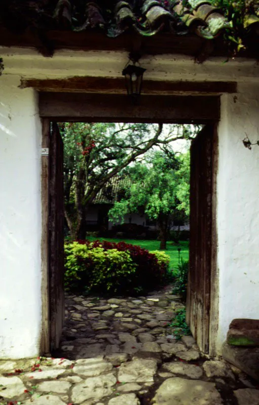 Baza, Valle de Tenza, Boyacá.
Una casa que “deja existir” el campo en torno suyo, a la manera de las alquerías árabes andaluzas. Nótese, abajo a la izquierda, la variante usual en Boyacá de la armadura de cubiertas en “par y nudillo” técnicamente primitiva, realizada usando maderas rollizas excesivamente delgadas a modo de pares. Estas, muy flexibles pero livianas
y baratas, se curvan bajo el peso del
tejado, y requieren riostra o jabalcones (más rollizas) apoyados en los tirantes, para sostenerlas. Tan confuso sistema artesanal refleja lo pintoresco de la ignorancia técnica
y lo divertido de
la improvisación.
A la derecha, el
acceso al patio es
un indudable acierto arquitectónico, reflejo del arte de saber entrar a los lugares.