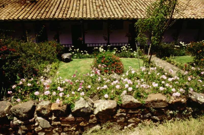 Casa de Suescún, Tibasosa, alrededores de Sogamoso, Boyacá. Localizada en lo que fue hace algunas décadas un lugar paradisíaco del valle
de Sogamoso. La hacienda misma, resultado del fraccionamiento,
en la primera mitad del siglo XVII, de
una vasta encomienda que abarcaba la comarca circundante, fue paulatinamente reducida hasta las escasas hectáreas que aún conforman el entorno de la casa.
El uso turístico
la ha salvado de
la desaparición o
la desfiguración gradual, pero hoy el lugar está sujeto a todas las amenazas propias del “desarrollo” económico e industrial: contaminación atmosférica, vecindad de urbanizaciones “pobres”, de un cementerio popular, etc. Resulta así milagroso que
Suescún haya conservado parte de
su grato ambiente
y la recóndita arquitectura de la casa.
Suescún no ofrece una arquitectura espectacular o efectista. La casa se limita a estar en su lugar de modo sencillo e íntimo. Se asoma modestamente sobre un leve altozano para observar el valle por sobre muros que, en gran parte, sólo existen excepto en el recuerdo.