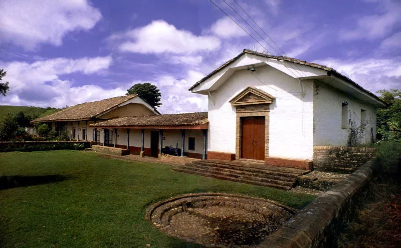 , Casa de
Antón Moreno,
Popayán, Cauca.
La más bella e inspirada relación de casa y paisaje entre las haciendas próximas a Popayán es sin duda la de Antón Moreno. La singular mezcla de dureza y suavidad de la arquitectura de la casa es la que presenta el paisaje circundante, y la una surge y depende de lo otro. Alcanzó su forma durante la última década del siglo XVIII, luego de terremotos y reformas sin cuenta, aunque la hacienda existía como “de trapiche” al final del siglo anterior, pasando luego a ser ganadería.
Antón Moreno es una síntesis de los esquemas básicos de ordenación espacial más usuales en la arquitectura de las casas de hacienda neogranadinas. Combina con elegancia un patio principal, atravesado por una rama sobre arquillos del complejo acueducto
–de raigambre islámica– de la casa, con galerías exteriores abiertas al panorama circundante. Incluye una capilla anexa ligada a la casa por el tramo que alberga las dependencias de servicios. Para estar a tono con la modestia arquitectónica de su espacio, la capilla posee un singular retablo pintado en falso relieve sobre lienzo.