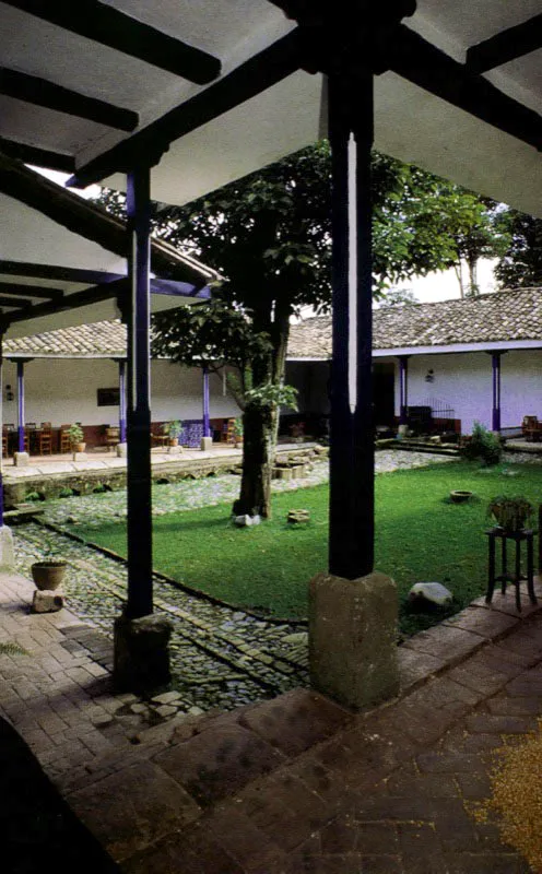 , Casa de
Antón Moreno,
Popayán, Cauca.
La más bella e inspirada relación de casa y paisaje entre las haciendas próximas a Popayán es sin duda la de Antón Moreno. La singular mezcla de dureza y suavidad de la arquitectura de la casa es la que presenta el paisaje circundante, y la una surge y depende de lo otro. Alcanzó su forma durante la última década del siglo XVIII, luego de terremotos y reformas sin cuenta, aunque la hacienda existía como “de trapiche” al final del siglo anterior, pasando luego a ser ganadería.
Antón Moreno es una síntesis de los esquemas básicos de ordenación espacial más usuales en la arquitectura de las casas de hacienda neogranadinas. Combina con elegancia un patio principal, atravesado por una rama sobre arquillos del complejo acueducto
–de raigambre islámica– de la casa, con galerías exteriores abiertas al panorama circundante. Incluye una capilla anexa ligada a la casa por el tramo que alberga las dependencias de servicios. Para estar a tono con la modestia arquitectónica de su espacio, la capilla posee un singular retablo pintado en falso relieve sobre lienzo.