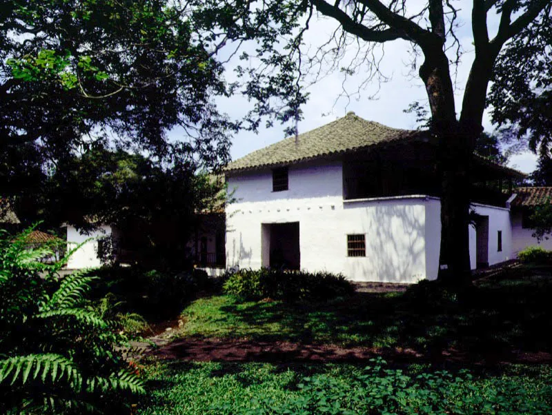 Piedechinche, El Cerrito, Valle del Cauca.
(Actual Museo de la Caña).
Las haciendas del Valle del Cauca fueron divididas en dos géneros, ya bien entrada la Colonia, para la implantación del cultivo de la caña de azúcar asiática en la región. Las haciendas productoras de azúcar pronto superaron en rendimiento a las de ganado vacuno. Algunas de las más prominentes haciendas de trapiche de la región, como El Alisal, han sido destruidas recientemente. Piedechinche pasó a una existencia, preferible a la desfiguración modernizante o la demolición, como museo histórico de
una actividad agrícola regional, con el mérito de hacer visible la organización espacial de una industria adscrita a una casa de hacienda.
Nótese cómo la casa
de los señores de Piedechinche ofrece similaridades espaciales con haciendas caucanas como Coconuco, Pisojé o Yambitará. Tales como la sobre-elevación de un tramo a manera de mirador y el uso de galerías exteriores de gran amplitud en piso alto y bajo, sin abarcar la totalidad de las fachadas.