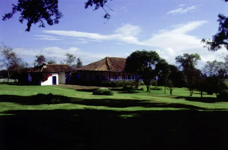 El Chacal, Tenjo, Cundinamarca.
Es una subdivisión, ocurrida al terminar el siglo XVII, de la enorme propiedad de Tibabuyes, en el occidente de la sabana de Bogotá y los alrededores de la población de Tenjo, Cundinamarca. La casa ilustrada aquí pudo haber sido construida inmediatamente luego de la configuración de la hacienda, es decir, al comenzar el siglo XVIII. A su vez, El Chacal fue subdividida en la segunda mitad del siglo XIX, por lo que cabe suponer que la “republicanización” decorativa de la casa colonial tuvo lugar en las últimas década de aquél.
La casa de El Chacal retiene su bella localización, dominada por la silueta de sus tejados, pero desprovista ya de la mayoría de las cercas y tapias circundantes, es decir, de los límites de espacios complementarios dentro de los cuales se inscribe su arquitectura.