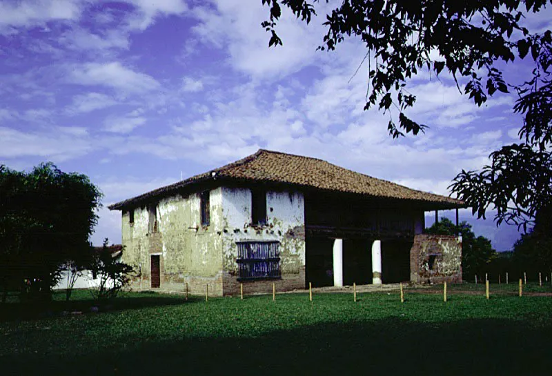 El Hato, Florida, Valle del Cauca.
Luego de la superposición de elementos eclécticos sobre la construcción de época colonial,
el siguiente paso en
la transición de las
casas de hacienda coloniales a las del período republicano fue la combinación de las tradiciones constructivas hispánicas con las nuevas modalidades compositivas para crear así edificaciones nuevas.
Un notable ejemplo de ese proceso es la casa de El Hato, cuya airosa volumetría establece una modalidad distinta en la relación entre casa y lugar. Sus rigurosas fachadas simétricas, construidas “a mano alzada”, le permitirían estar situada indistintamente en cualquier lugar de la región (lo cual no se podría decir, por ejemplo, de La Concepción de Amaime, o La Merced). El Hato inaugura, en cierto modo, la época de la casa de hacienda vallecaucana moderna, abstracta, independiente de su propia localización, aunque sin renegar por ello de su pasado colonial. Nótese en esta y otras casas de la época la combinación muy original de tejados a la manera campesina y, bajo éstos, fachadas academizantes.