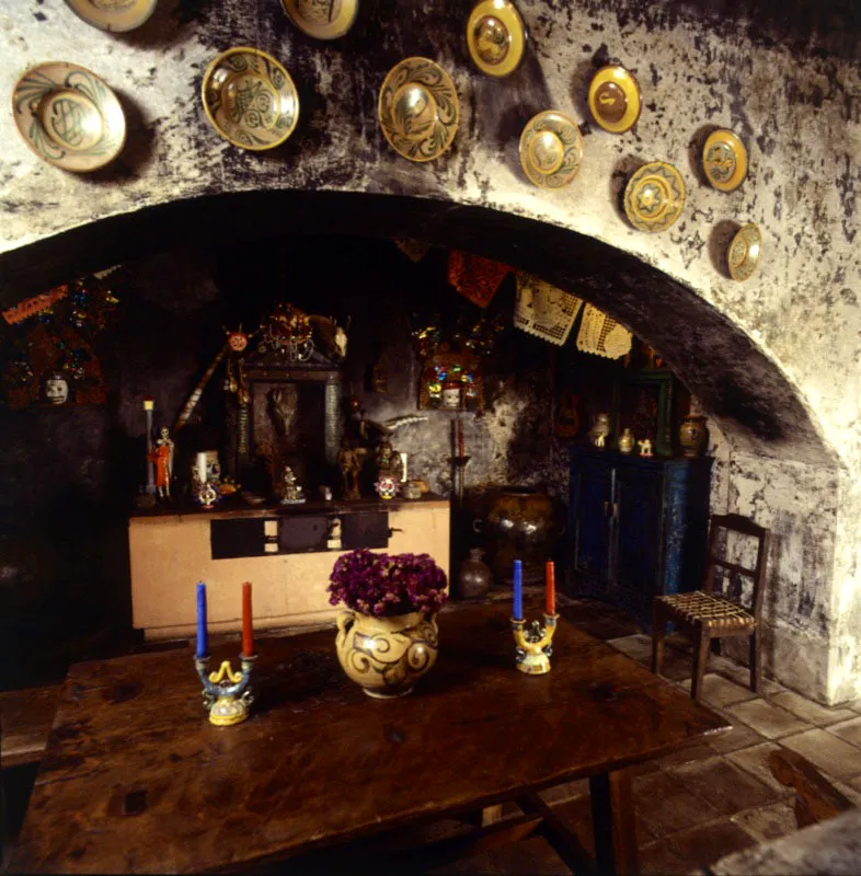 Parte de la antigua cocina de la casa, en la que se aprecia el arco rebajado de la chimenea adornada con platos de maylica, una mesa antigua en primer plano y, al fondo, sobre la vieja estufa, un conjunto de objetos y figuras de santos a manera de altar popular. 