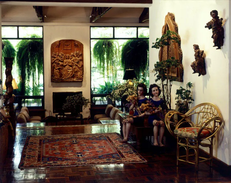 Corredor de una casa contemporánea de la capital, con grandes ventanales al fondo. Sobre la mesa y la pared imágenes coloniales, talladas y policromadas.