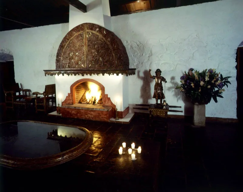 Gran salón, dominado por una chimenea con relieve tallado sobrepuesto, varios muebles antiguos e imágenes que resaltan los muros rústicos con su encalado tradicional.