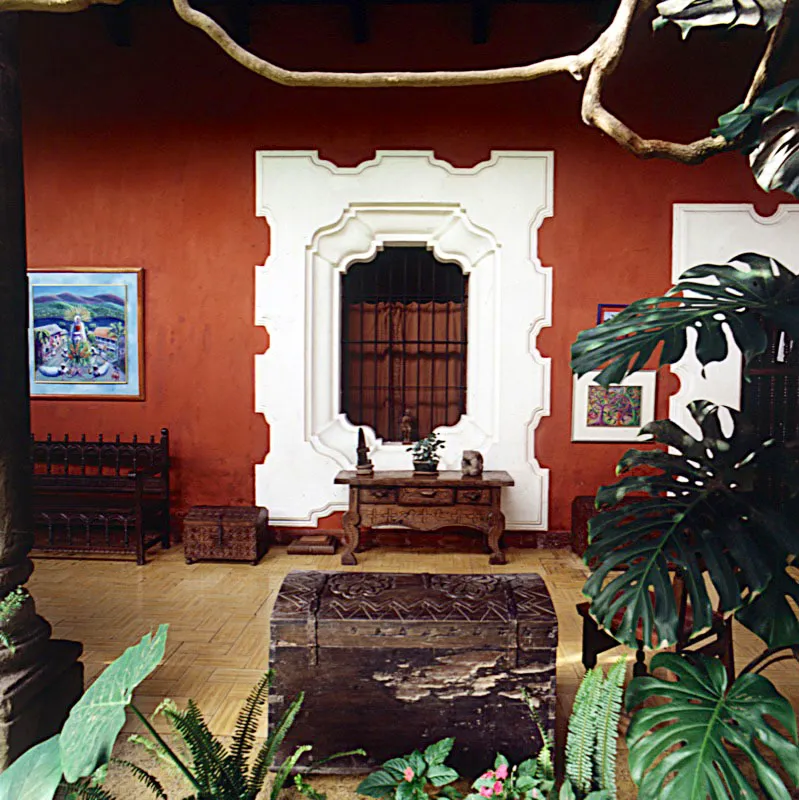 Ventana colonial con su marco encalado blanco sobre el muro rojo profundo, un óleo de la pintora Deborah Duflon, a la izquierda, y otras piezas de mobiliario rústico.