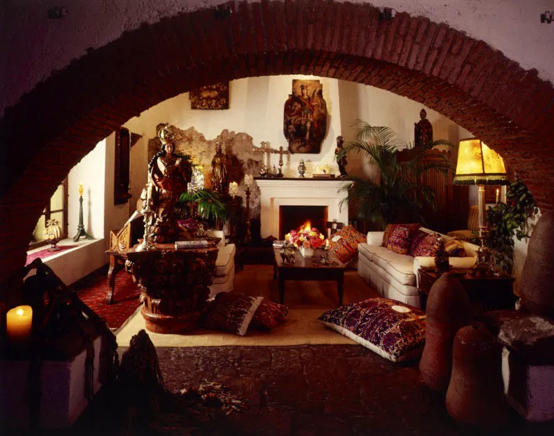En esta sala antigüeña, que observamos a través del arco rebajado de ladrillo visto, se aprecian varias esculturas y piezas coloniales. La primera, una Virgen con corona de plata sobre gran capitel de madera; al fondo, donde se combina el revoque con una parte de la pared antigua, otras esculturas; sobre la chimenea, en cuya repisa hay varias piezas de plata, la pintura de un arcángel; y a la derecha un armario tradicional. Los sofás modernos en tapicería blanca tienen varios cojines recubiertos con telas indígenas en los que predominan el rojo y el morado.