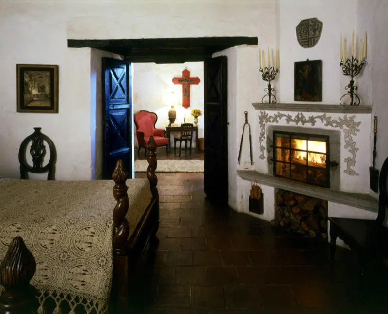 Dormitorio antigüeño de casa restaurada, con chimenea contemporánea adaptada, que se comunica con un pequeño
salón a través de una puerta sobre grueso muro colonial. La cruz que se observa al fondo, sobre el bastidor de madera roja, también es colonial.