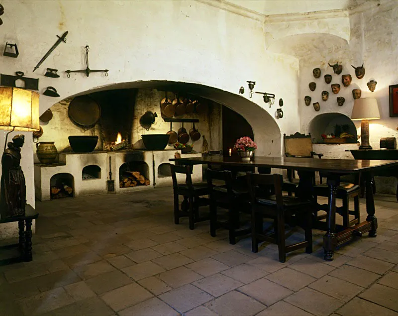 Antigua cocina colonial transformada en comedor, decorado con diversos objetos de hierro, máscaras de madera, ollas de bronce, lámparas con base de figura de santo, y muebles tradicionales.