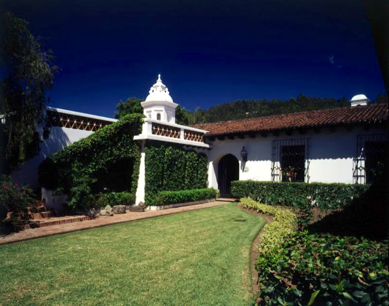 Acceso a casa contemporánea de Antigua y vista lateral a través del jardín. Se combinan aquí la arquitectura tradicional y una amplia zona verde que aprovecha, entre la vegetación, vestigios coloniales de piedra procedentes de antiguas edificaciones destruidas en el terremoto de 1773. Al lado de la casa, una amplia entrada empedrada para carruajes y una pila rodeada de buganvillas.
