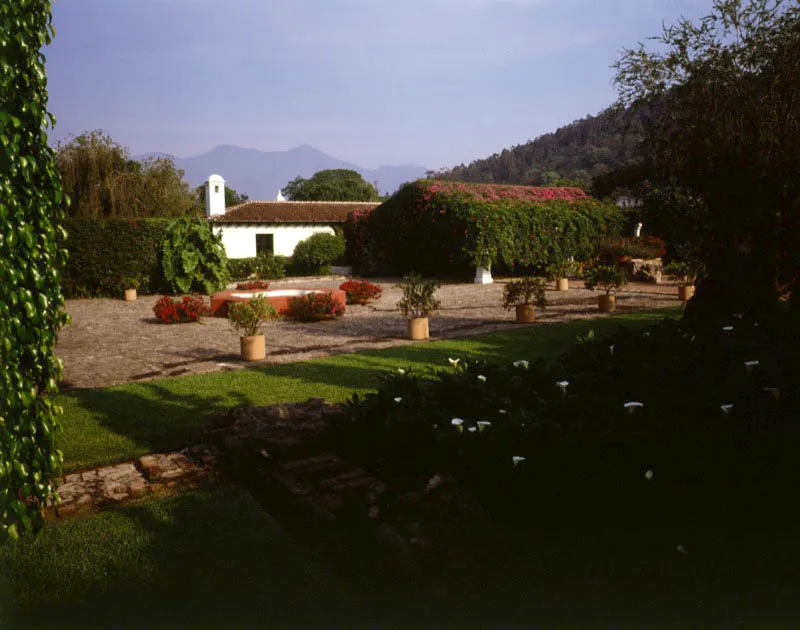 Acceso a casa contemporánea de Antigua y vista lateral a través del jardín. Se combinan aquí la arquitectura tradicional y una amplia zona verde que aprovecha, entre la vegetación, vestigios coloniales de piedra procedentes de antiguas edificaciones destruidas en el terremoto de 1773. Al lado de la casa, una amplia entrada empedrada para carruajes y una pila rodeada de buganvillas.