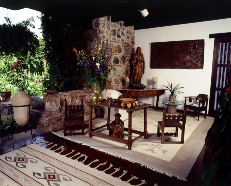 Al final de un largo corredor abierto hacia el jardín, este pequeño ambiente decorado con un dhurry de la India en tonos suaves. Un conjunto de sillas rústicas, que forman un área de estar, y dos tallas de madera del siglo XVIII complementan el rincón. Al fondo la pared antigua sin repello, compuesta de diferentes tipos de piedra y un gran retablo tallado de madera.