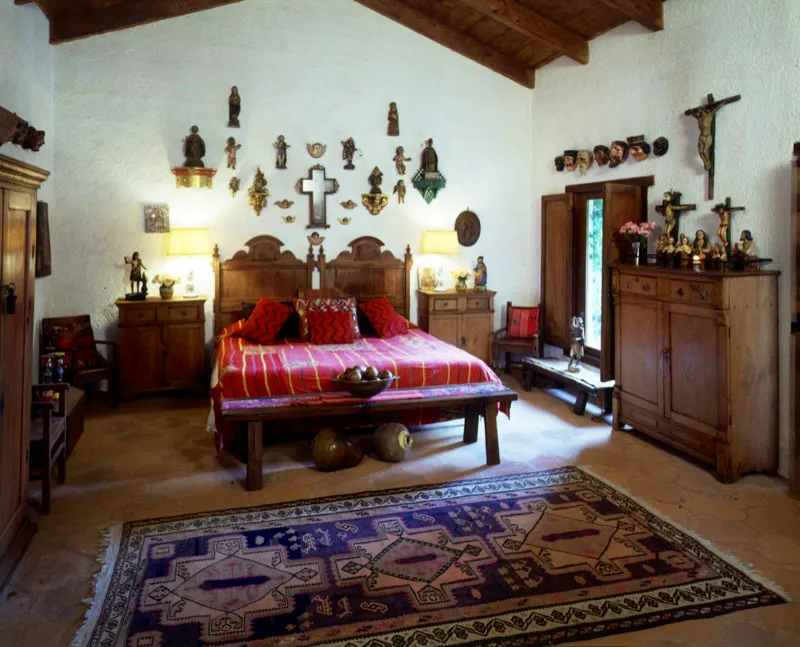 Otro dormitorio de la misma casa, con similar concepto decorativo, donde abundan, tanto en la pared como sobre los muebles, pequeñas esculturas coloniales, populares y cultas, entre las que hay varios crucifijos y máscaras de este siglo. El cubrecama y los cojines son de textiles guatemaltecos indígenas.