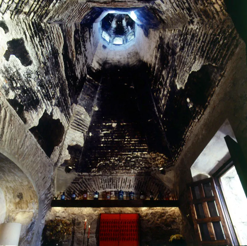 Dos vistas del mismo ambiente en una casa de Antigua, restaurada por el ingeniero guatemalteco Amerigo Giracca. Se trata de una vieja cocina que conserva su chimenea acupulada en ladrillo. En la restauración se conservó incluso el ahumado y se adaptó a su nuevo uso como sala, con muebles tradicionales y modernos, una repisa con frascos de farmacia antigua y, en la parte inferior, un huipil maya de vivo color rojo, enmarcado.