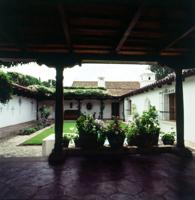 Casa antigüeña reconstruida. En primer plano, una galería con baldosa tradicional y plantas. A la izquierda del patio engramado, un corredor con cruces de hierro forjado en el muro. Al costado derecho, así como al fondo, se han reproducido las típicas ventanas coloniales lobuladas.