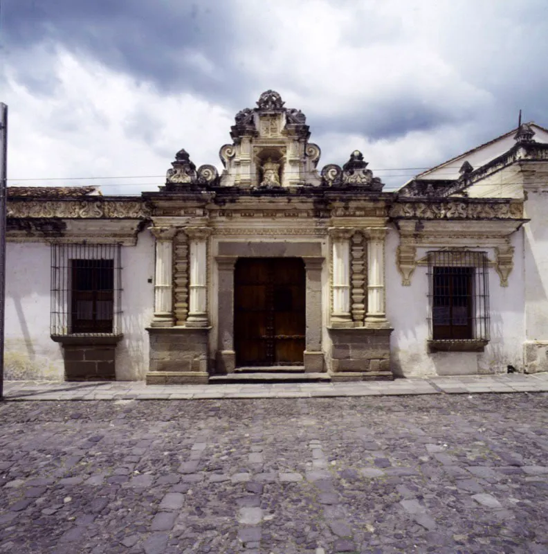 La elaborada portada principal del mismo edificio, en la que se combinan elementos tpicos del ultrabarroco en Santiago de Guatemala: columnas revestidas de pilastrillas y en medio pilastras almohadilladas; en el entablamento diversos ejemplos de ornamentacin de la poca; y arriba una hornacina enmarcada por roleos y pilastrillas, en cuyo interior se encuentra la Virgen de la Asuncin, en argamasa.  