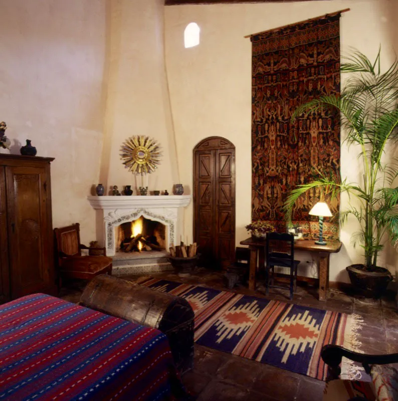 Dormitorio de la misma casa antigüeña, con chimenea adornada con incrustaciones de azulejo y coronada con un espejo de resplandor dorado. A la izquierda, el armario primitivo de madera se complementa con la alfombra de tejido indígena. De la pared cuelga un textil ikat de Indonesia.