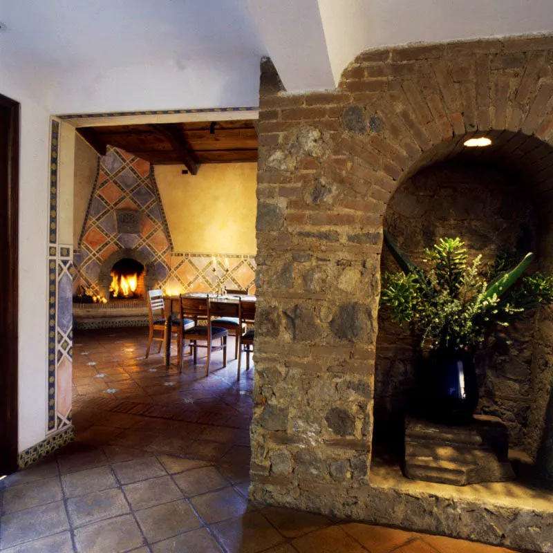 Acceso al comedor de una casa en Antigua, precedido por un nicho sobre muro de piedra y ladrillo a la vista, con chimenea contemporánea policromada a mano y boca en arco de piedra. El piso es de baldosa de barro salpicado de azulejos.
