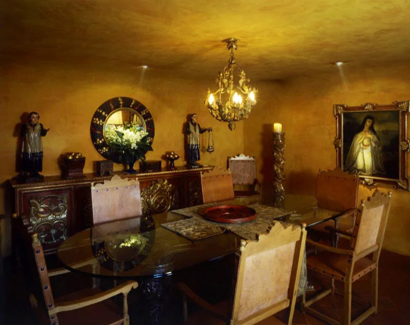 En Antigua, comedor dentro de espacio restaurado, con paredes de esquinas redondeadas, estucadas, mesa contemporánea de vidrio y sillas de estilo español. A la derecha, pintura colonial y columna salomónica en madera tallada. Al fondo, aparador con relieves en pan de oro que sostiene dos monaguillos coloniales de madera policromada. Al centro, reproducción de espejo redondo colonial.