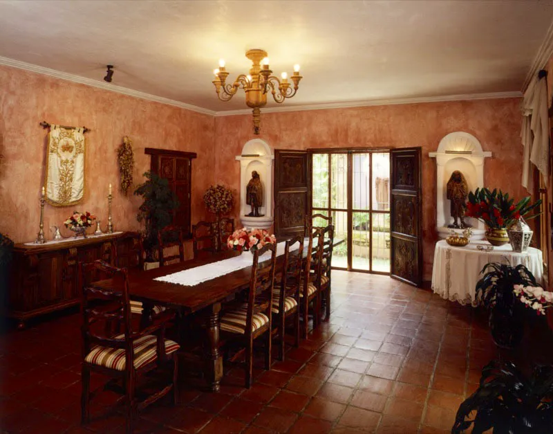 Comedor de la casa anterior. Al centro, sobre una mesa contempornea, pende una araa de madera y hierro forjado.Al fondo, dos hornacinas con imgenes de madera tallada y a la izquierda una casulla colonial colgada del muro sobre un trinchante. Piso de baldosa de terracota y puertas antiguas talladas, incorporadas a la estructura.
 