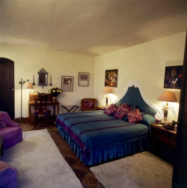 Dormitorio principal con cama cubierta y espaldar tapizado, y cojines forrados con textiles típicos guatemaltecos.
Al fondo, huipiles antiguos enmarcados y espejo de estaño labrado. Al lado pinturas primitivas y lámparas fabricadas utilizando ángeles de madera como soporte.