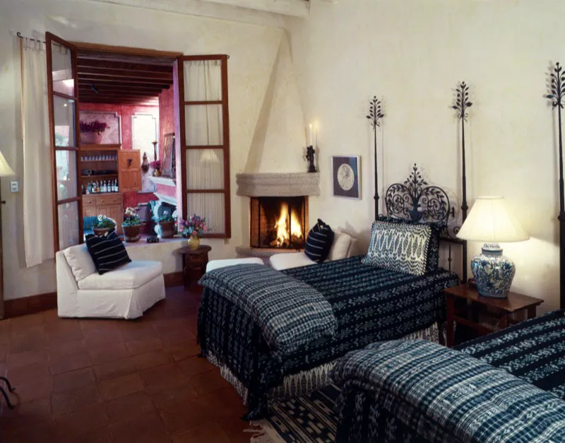 Habitación de la misma casa con piso de baldosa de barro y chimenea contemporánea en la esquina. Las camas con espaldar de hierro forjado están cubiertas con textiles guatemaltecos con motivos de jaspe.