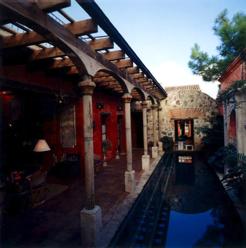 Pequeño salón abierto a un corredor flanqueado por una piscina longitudinal, paralela al muro del fondo, con bocas de agua y jardín en los nichos. Los muebles son equipales cubiertos por cojines tejidos y mesas auxiliares en forma de tambor. La imagen de arriba muestra la longitud de la piscina, las columnas tradicionales en madera rolliza y el rústico muro de piedra al fondo.