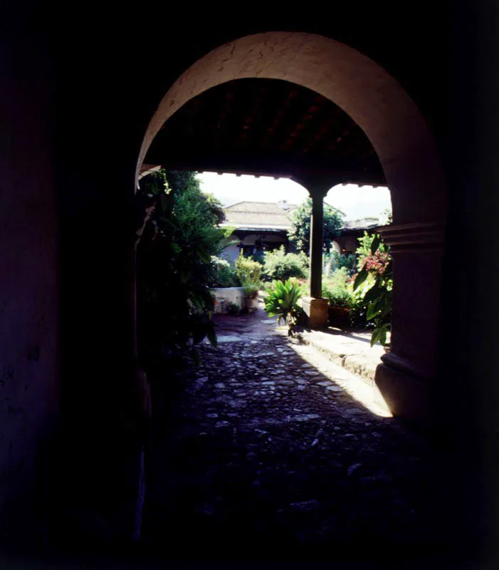 Zaguán empedrado de entrada a casa de Antigua, a través de un arco. Se aprecia el patio jardinizado, el escalón que sube al corredor, las columnas cilíndricas y las zapatas tradicionales.