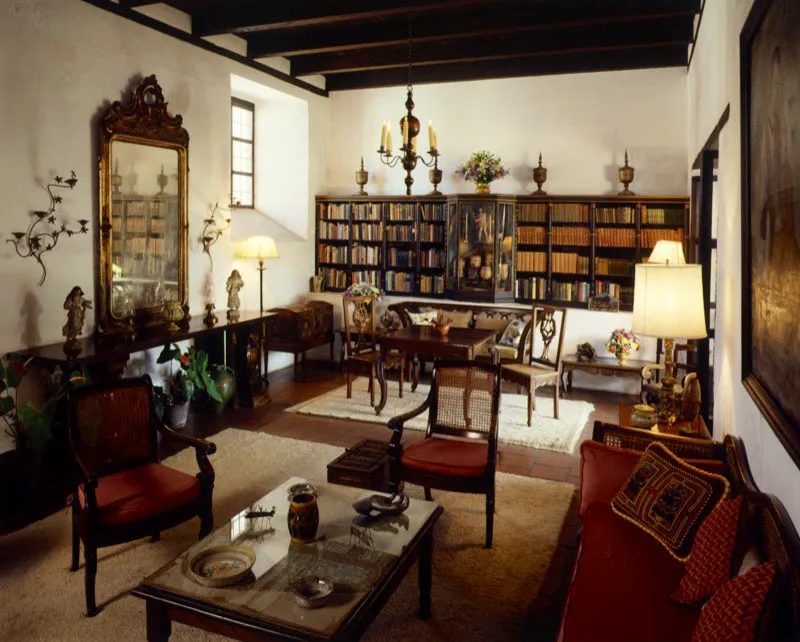 Saln y biblioteca de la misma casa en Antigua. El mobiliario y la decoracin son muy variados y conforman un conjunto agradable. A la izquierda, sobre el aparador, un monumental espejo tallado y dorado, flanqueado por candelabros de hierro forjado contemporneos. Al fondo, una biblioteca con vitrina central, que contiene piezas mayas y tallas coloniales, y a la derecha un gran leo colonial.
 