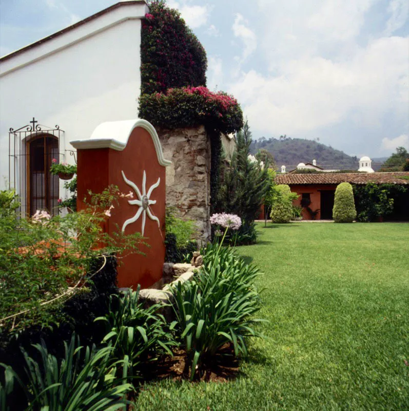 Casa contemporánea en el marco de la ciudad de Antigua. En un amplio sitio se ha combinado un hermoso jardín con arquitectura contemporánea que incorpora ruinas de piedra colonial. Una pequeña fuente contemporánea decorada con un sol en relieve, un búcaro en mayólica local y restos respetados de construcción antigua, cubiertos en parte por una buganvilla.