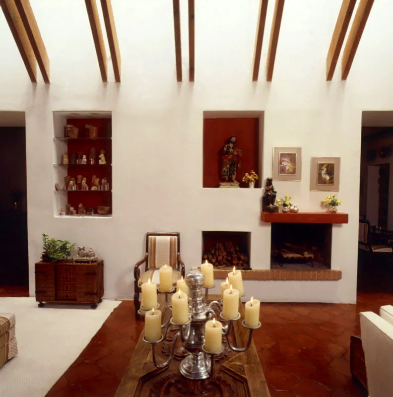 Muro del gran salón de la casa antigüeña anterior. Predomina la línea recta, el color blanco y los espacios cuadrados. En los nichos, donde el color zapote combina con la baldosa, se exhiben objetos prehispánicos y una escultura colonial en madera policromada. En primer plano, una mesa hecha de una puerta antigua, con un interesante candelabro de estaño de diez brazos. Sobre la chimenea, dos obras del pintor guatemalteco Roberto González Goyri.