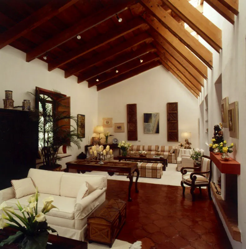 Amplio salón en tonos blancos y terracotas. Se observan piezas coloniales de madera, como el baúl tallado junto al sofá blanco, la mesa rectangular que divide los ambientes, la esquinera con el atril colonial de plata y las dos puertas talladas sobre el fondo, que enmarcan un cuadro del pintor de origen cubano Óscar Magnan. A la izquierda, dos témperas del guatemalteco Carlos Mérida y al lado de la ventana un óleo de la pintora Irma Luján, también guatemalteca.