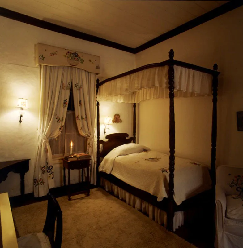 Casa en Cobán, Alta Verapaz. Un dormitorio en el que predomina el blanco, tanto en las paredes, como en las cortinas, los muebles, la alfombra y la cama. Esta última, con baldaquino, tiene un cubrecama bordado
con flores de colores, al igual que la cortina.