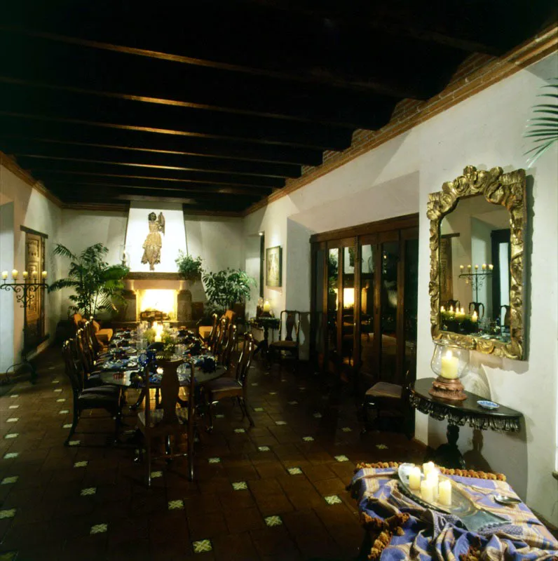 Amplio comedor con baldosa de barro y pequeños azulejos intercalados, en la misma casa reconstruida de Antigua. Sus dimensiones, el color blanco de los muros y las vigas del techo le confieren un carácter austero.
Al fondo, sobre la chimenea, un gran arcángel en relieve de madera policromada, frente a una pequeña sala de estar. A la derecha, espejo barroco dorado en pan de oro y consola de madera tallada.