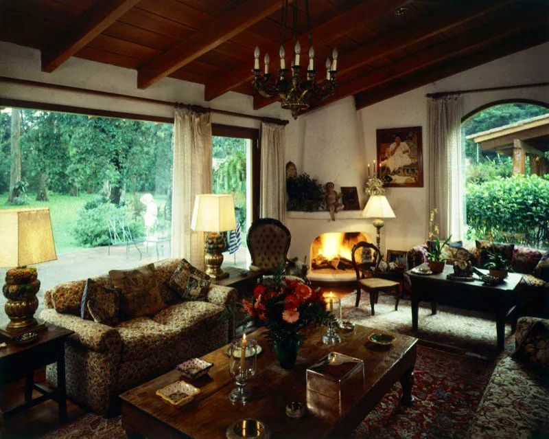 Esta sala de amplias ventanas se abre a un bonito jardín en una casa de la ciudad de Guatemala. Se mezclan en ella elementos variados: sillones modernos, sillas tradicionales, mesas rústicas de madera, alfombras orientales, vigas de madera y una araña de bronce. Sobre las mesas sigue la variedad: lámparas modernas de madera tallada en forma de fruteros, piezas de plata, ceniceros de cerámica, un Niño Jesús sentado sobre la repisa de la chimenea, un candelero de plata, y un cuadro del artista español Miguel García Luque.