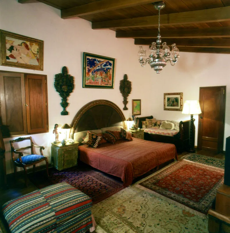 Dormitorio de la misma casa en ciudad de Guatemala. La cama, con cobertor guatemalteco y cojines de kilims, tiene un respaldar semicircular de madera, flanqueado por dos relieves tallados. A la derecha, un retrato realizado por el pintor guatemalteco Ramón Banus y arriba de la cama, dos óleos del español Miguel García Luque. Del techo pende una araña de estaño, y el piso de duela de madera está cubierto por alfombras orientales.