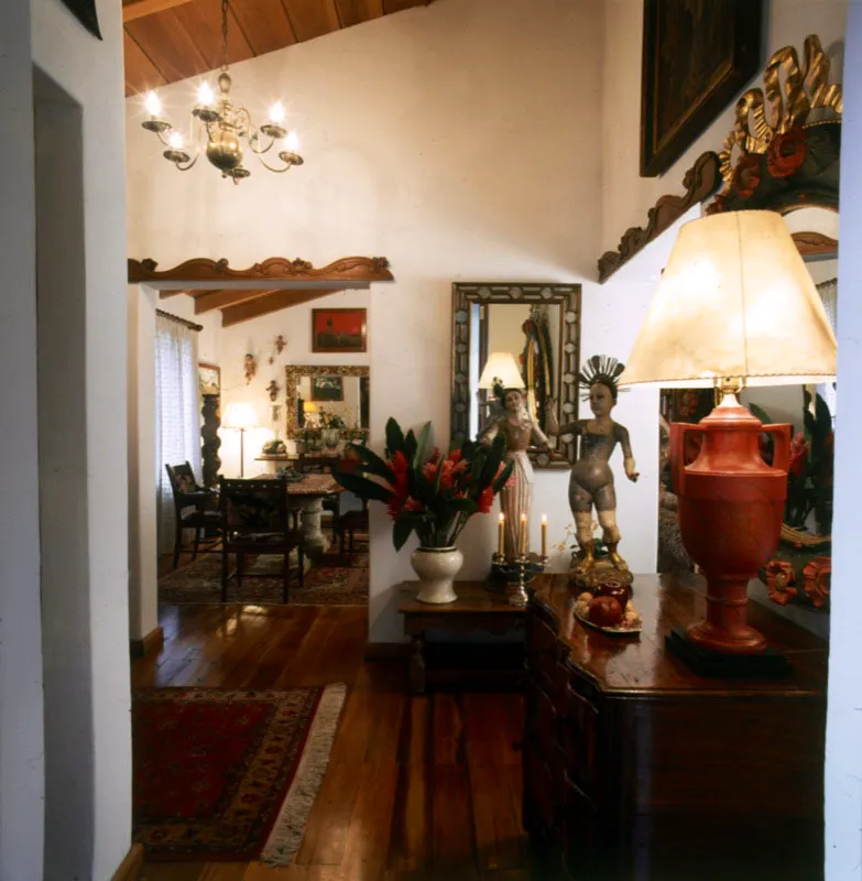 Vestíbulo de entrada al comedor, con santos de vestir coloniales, espejo de marco calado y roleos coloniales en madera tallada. Al fondo, en el comedor, se aprecian las bases de piedra de la mesa, una columna salomónica, pinturas de Miguel García Luque y varias imágenes del Niño Jesús.