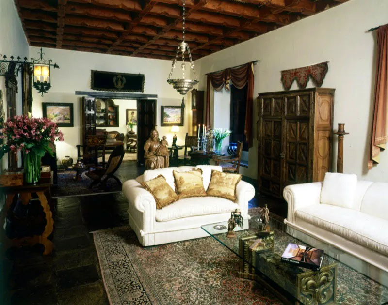 Perspectiva más completa de la sala de la página 178. Sobre la derecha puede verse un armario de madera tallada, y sobre la izquierda una consola colonial. En la moderna mesa de centro, en lámina de vidrio sobre bases de madera tallada y dorada, hay unas interesantes muestras de pequeñas esculturas coloniales guatemaltecas. Del artesonado cuelga una lámpara colonial en plata. Sobre la puerta del fondo, una pieza de terciopelo de la época colonial, y sobre el armario un textil antiguo bordado.