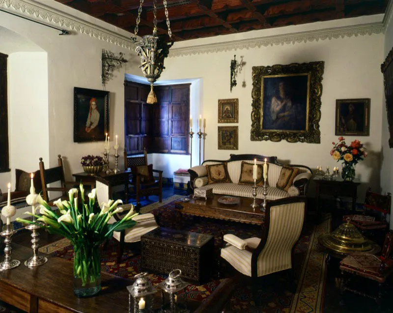 Otra perspectiva del salón mostrado en la página 179. Se observan distintos tipos de sillas y sillones republicanos; mesas de madera, de diferente procedencia, con candeleros de plata; así como un cofre español taraceado. En el muro del fondo, varias pinturas coloniales y, en el ángulo del fondo, las hojas cerradas de una ventana o balcón de esquina, una solución arquitectónica común a toda Centroamérica.