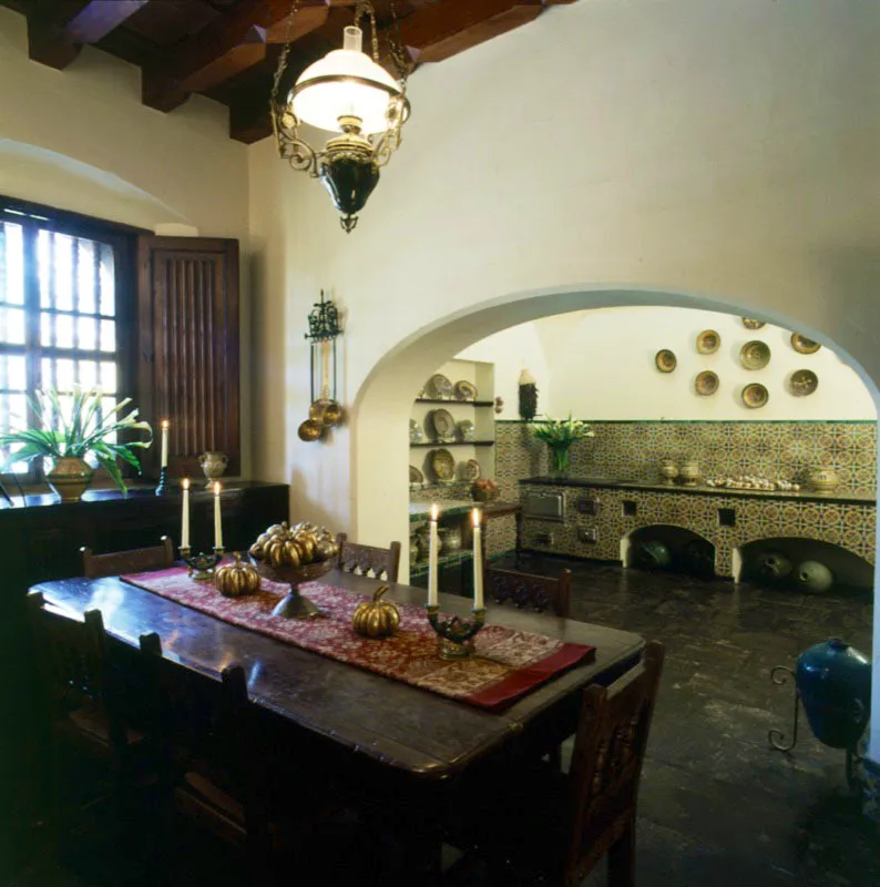 La cocina de la casa integrada, a través de un arco, con el desayunador. En primer plano, una mesa rústica con sillas, sobre la que aparecen dos candeleros de mayólica antigüeña y calabazas de madera dorada sobre un textil oriental. Detrás del arco rebajado, se aprecia la cocina restaurada, con profusión de azulejos y platos de mayólica local, tanto en la pared como en la alacena lateral.