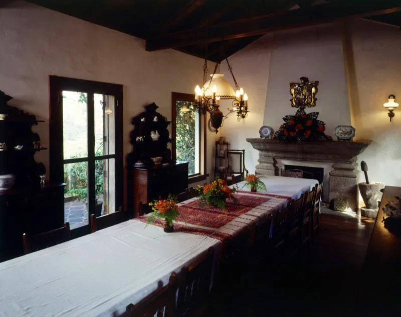 Detalle del comedor de otro ejemplo de casa patronal, en finca cercana a Antigua. En el centro, una mesa sencilla y larga, con sillas rústicas, mantel típico de varias piezas unidas y una parte bordada en el centro. A la izquierda, alacena de madera de la segunda mitad del siglo XIX. Sobre la chimenea en piedra tallada, dos platos de cerámica de tipo chino y arriba el escudo familiar tallado en madera con dorado y policromía. Puede verse parte de las vigas de madera de la techumbre de dos aguas, así como una lámpara eléctrica que imita una araña con velas.