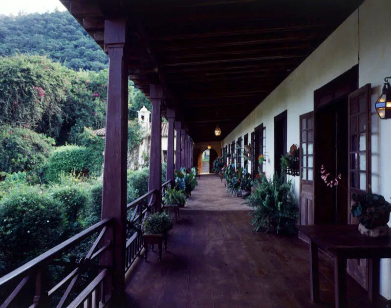 Vista del corredor de la misma casa, con terraza espaola sostenida por pilares de madera, sin muebles, adornado slo con macetas de plantas. El piso del acceso es en baldosas de barro, y su prolongacin en voladizo es en duela de madera, con baranda del mismo material.  