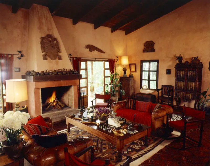 Casa de campo contemporánea en las afueras de la capital. La sala en color rosa está organizada alrededor de dos sofás con tapicería de cuero y textil sobrepuesto, cojines de tela típica y una mesa de centro, decorada con diferentes tallas de animales en madera, de creación popular, y otros objetos. Una gran chimenea, en cuya repisa de madera decorada con jarras de cerámica vidriada, pende una rústica talla de un águila bicéfala. Complementan el salón diversos muebles, con pequeñas esculturas y piezas de arte.