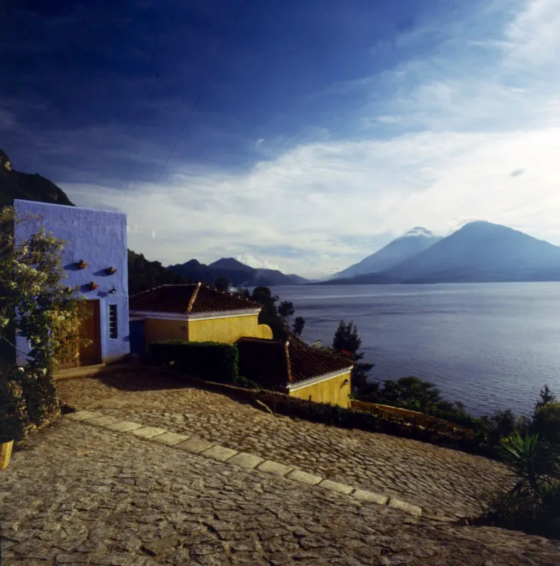 Casa de recreo ubicada en la parte alta del norte del lago de Atitlán, con una excelente vista de los volcanes Tolimán y Atitlán. Alrededor de este lago se han construido numerosas casas de recreo de muy diversos estilos. A la izquierda se ve el área de acceso a la casa, con terraplenes empedrados y muros de colores azul y amarillo.