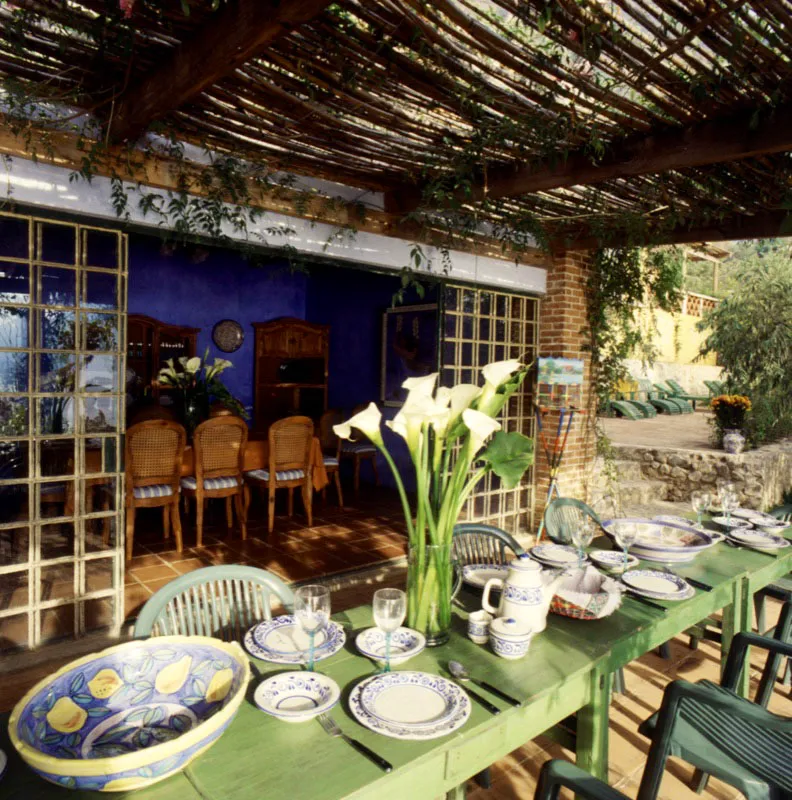 Comedores de la casa. En primer plano, el desayunador informal, en una terraza bajo un toldo de cañas y una enredadera; las mesas rústicas están pintadas de alegres colores y la vajilla es en cerámica de Antigua, pintada a mano. Al fondo se aprecia el comedor formal, con muros de color añil.