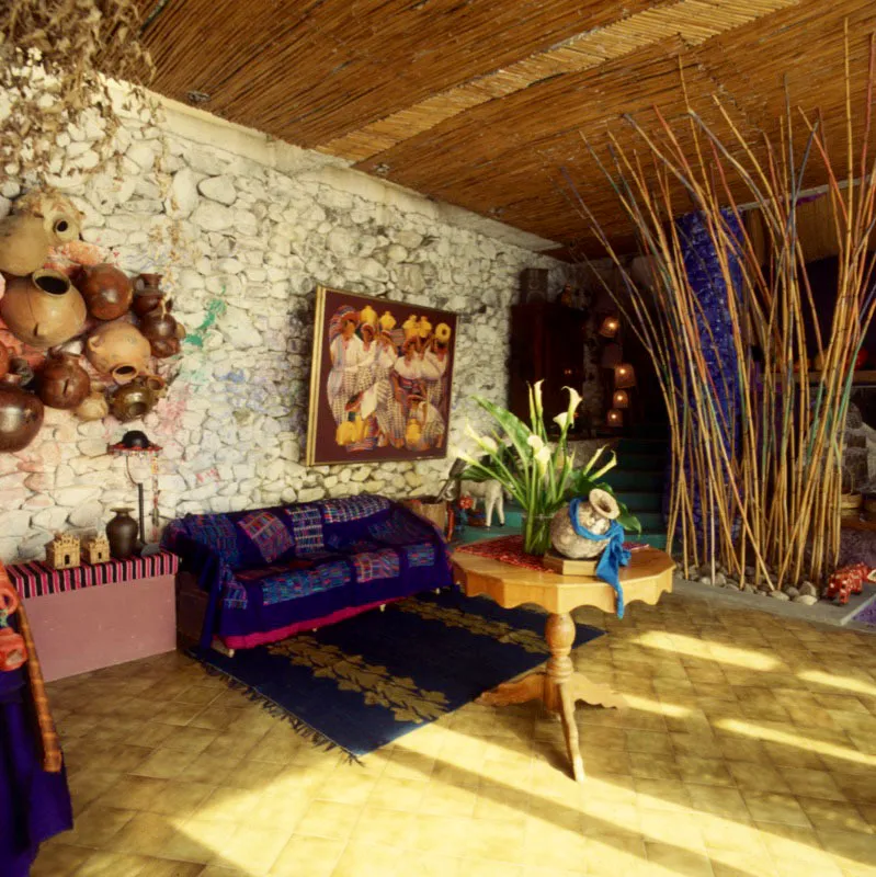 Ambiente interior de otra casa de recreo contemporánea a orillas del lago de Atitlán, con pared en piedra rústica, piso en adoquín de cemento pintado, decorado con alfombra de la zona de Momostenango, y textiles, de procedencia indígena, sobrepuestos en el sofá. A la derecha, un mortero de madera y, sobre el muro, un conjunto de garrafas de barro populares y una pintura de David Ordóñez. El cielo raso es en cañas de la región.