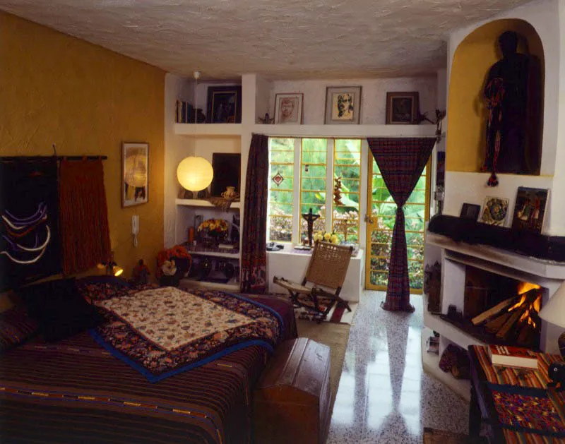 Dormitorio de la misma casa del artista, en ciudad de Guatemala. La cama tiene una colcha de tela indígena guatemalteca, cubierta por un textil oriental, y delante un baúl de madera sencillo. Al fondo, silla de tijera, tejida en fibra de agave, y a la derecha, chimenea con nicho que contiene un santo primitivo.