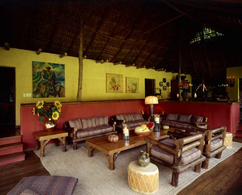 Sala con mostrador que sirve de bar. Los muebles en madera rolliza, equivalente a la estructura del ranchón, fueron fabricados en la zona y se complementan con el lugar. En el muro amarillo del fondo, se puede ver un cuadro de la primera época de Erwin Guillermo, y dos de Salvador Gálvez, ambos guatemaltecos.